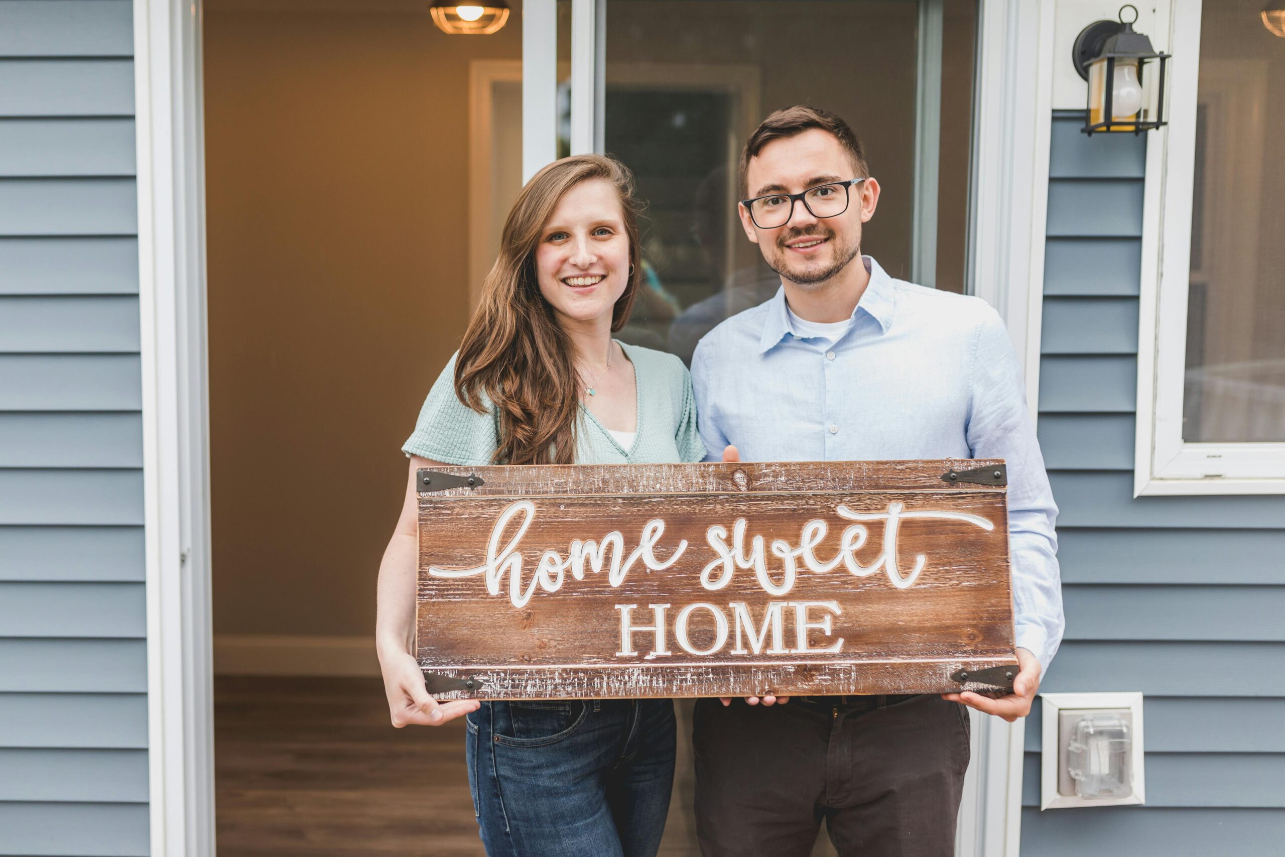 Smiling couple holding 'Home Sweet Home' sign in front of their new house entrance.